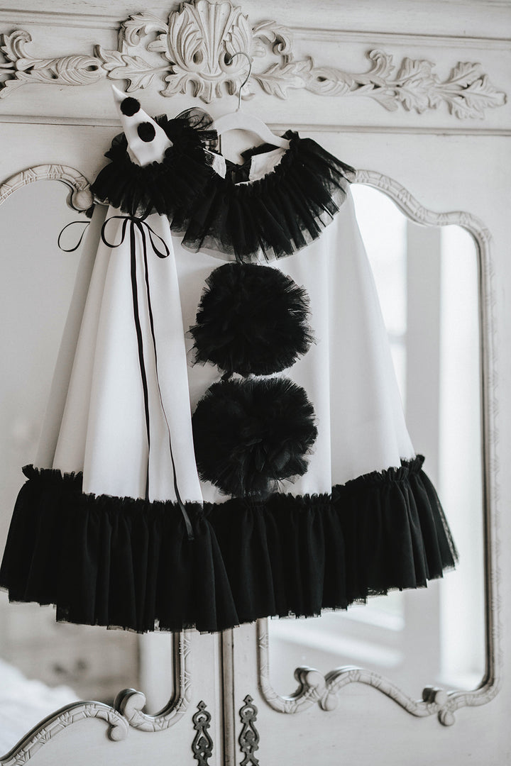 White satin Pirouette clown cape hanging on a closet with black tulle ruffle trim, oversized pom-poms and matching pointed hat.