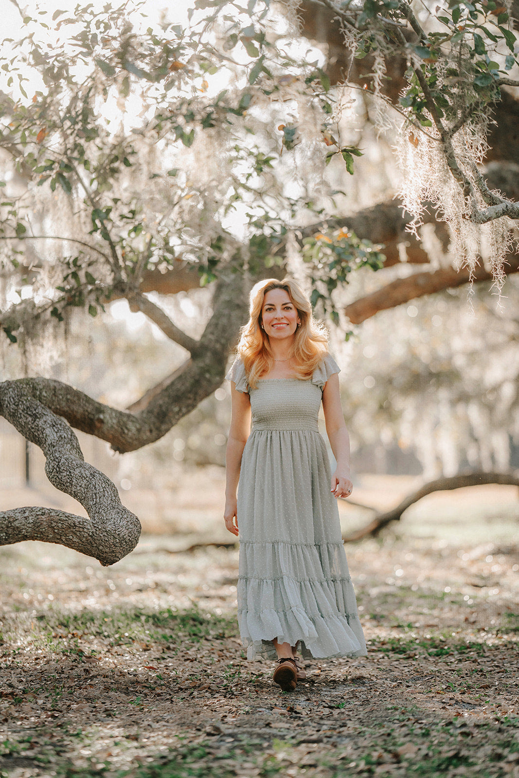 Woman walking through an orchard in the Bay Leaf sage green Swiss dot maxi dress with flutter sleeves.