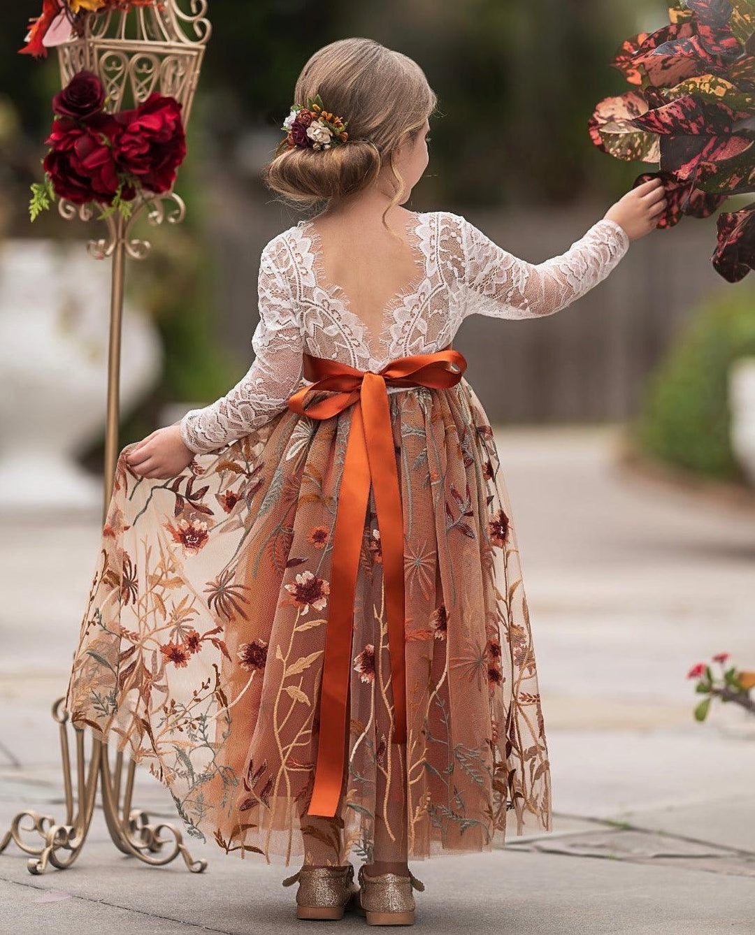Woman in a floral dress with an orange ribbon standing next to decorative floral arrangements.