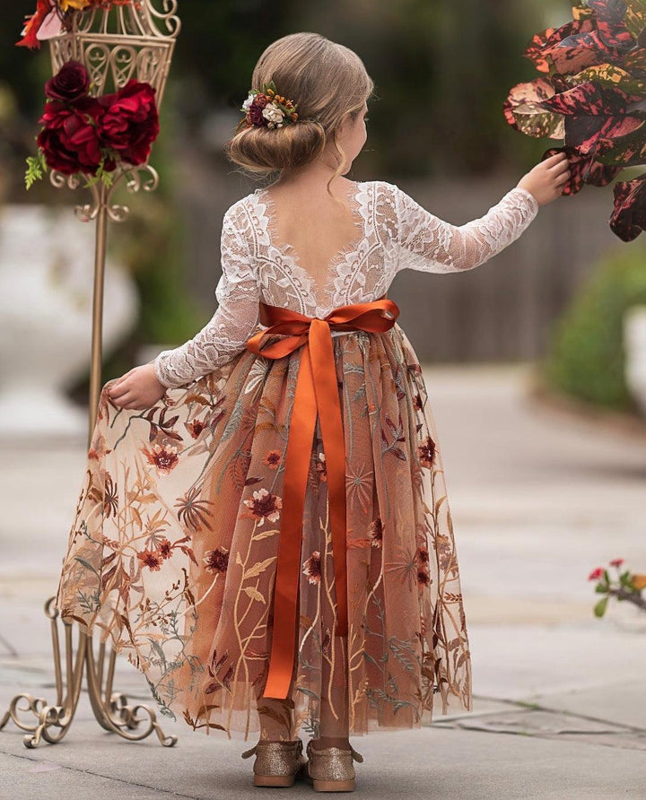 Woman in a floral dress with an orange ribbon standing next to decorative floral arrangements.