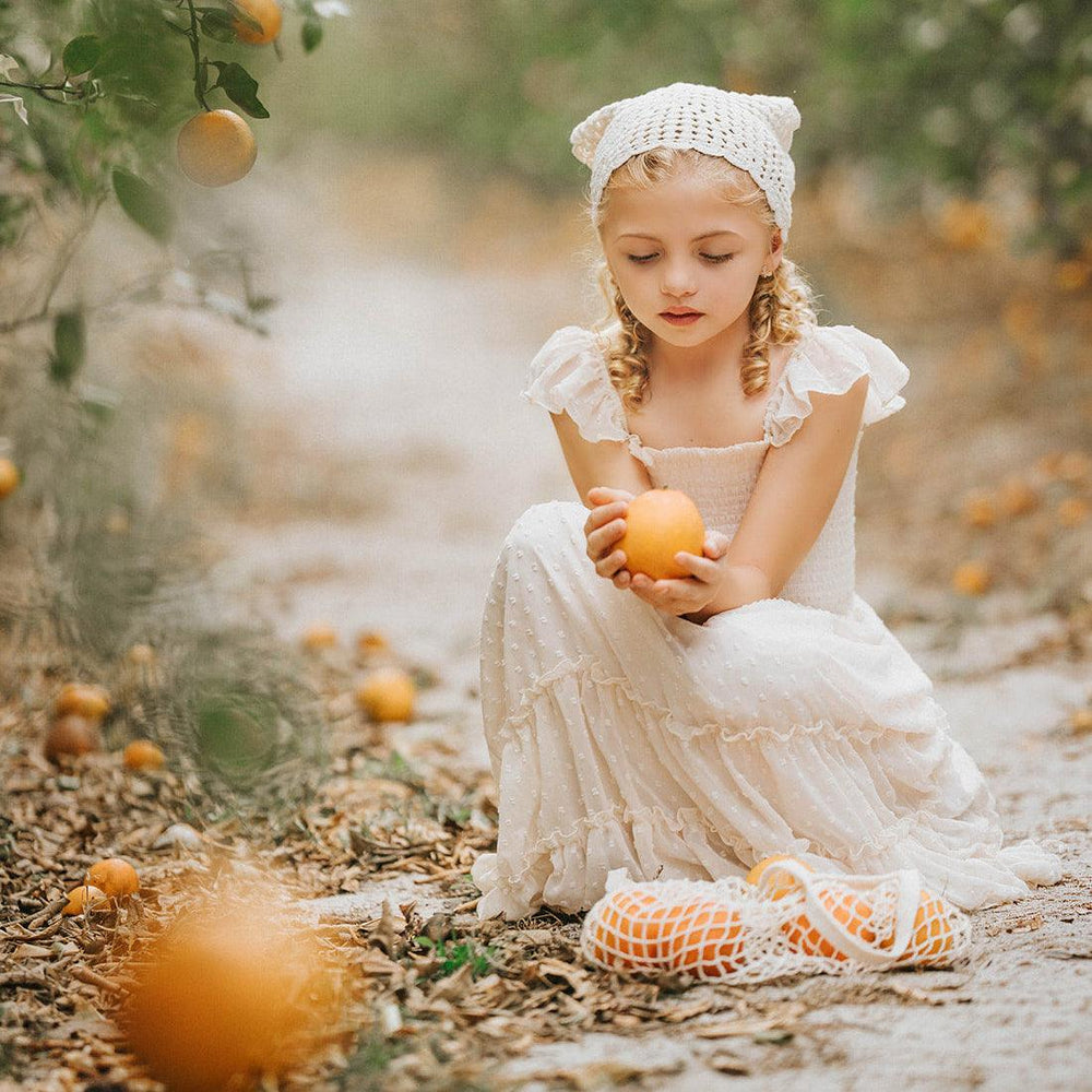 Child sitting in an orchard wearing the Citrus Cream Orchard Dress with fresh citrus fruit beside her