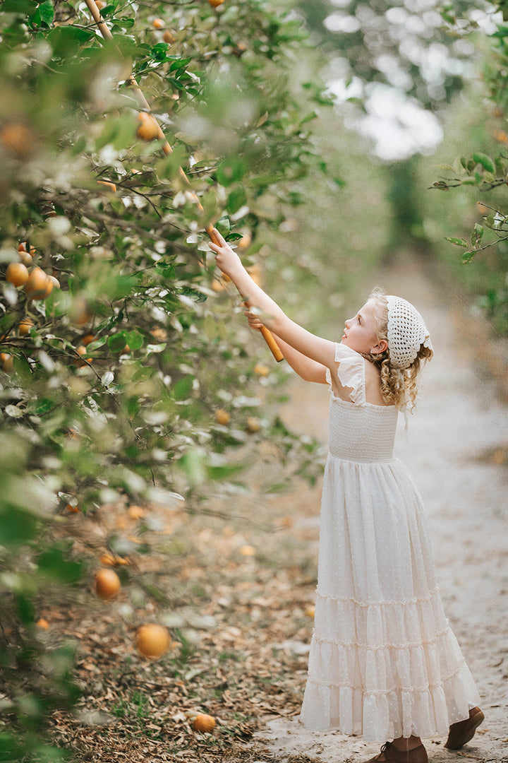 Girl reaching toward orchard branches while wearing the Citrus Cream Orchard Dress in warm natural light