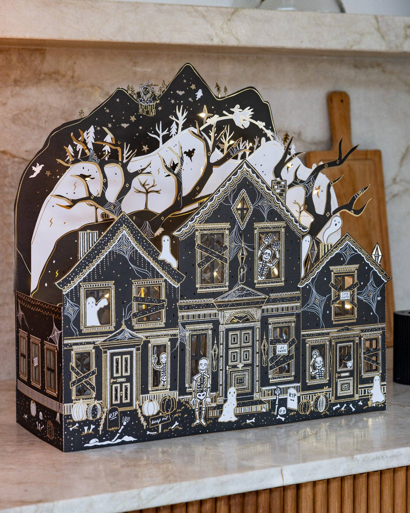 Storybook-inspired haunted paper house glowing with warm lights beside black taper candles for Halloween decorating.