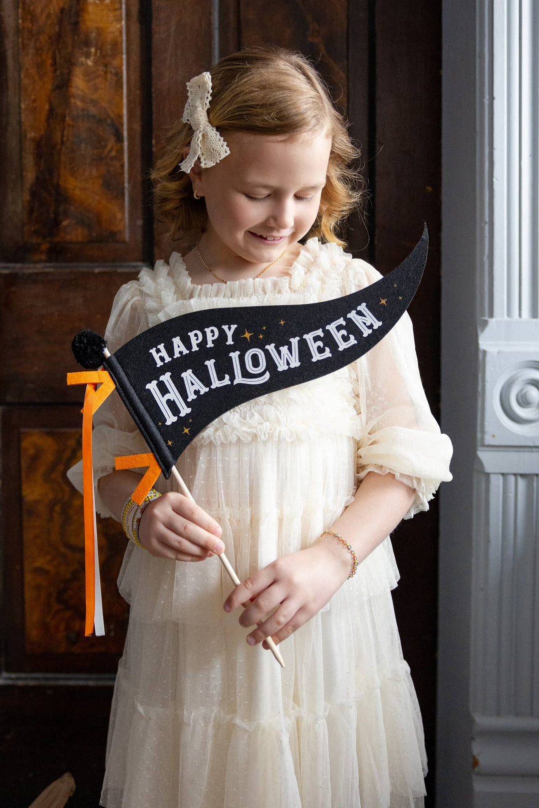 Little girl in a cream dress holding a black Happy Halloween felt pennant with orange ribbons