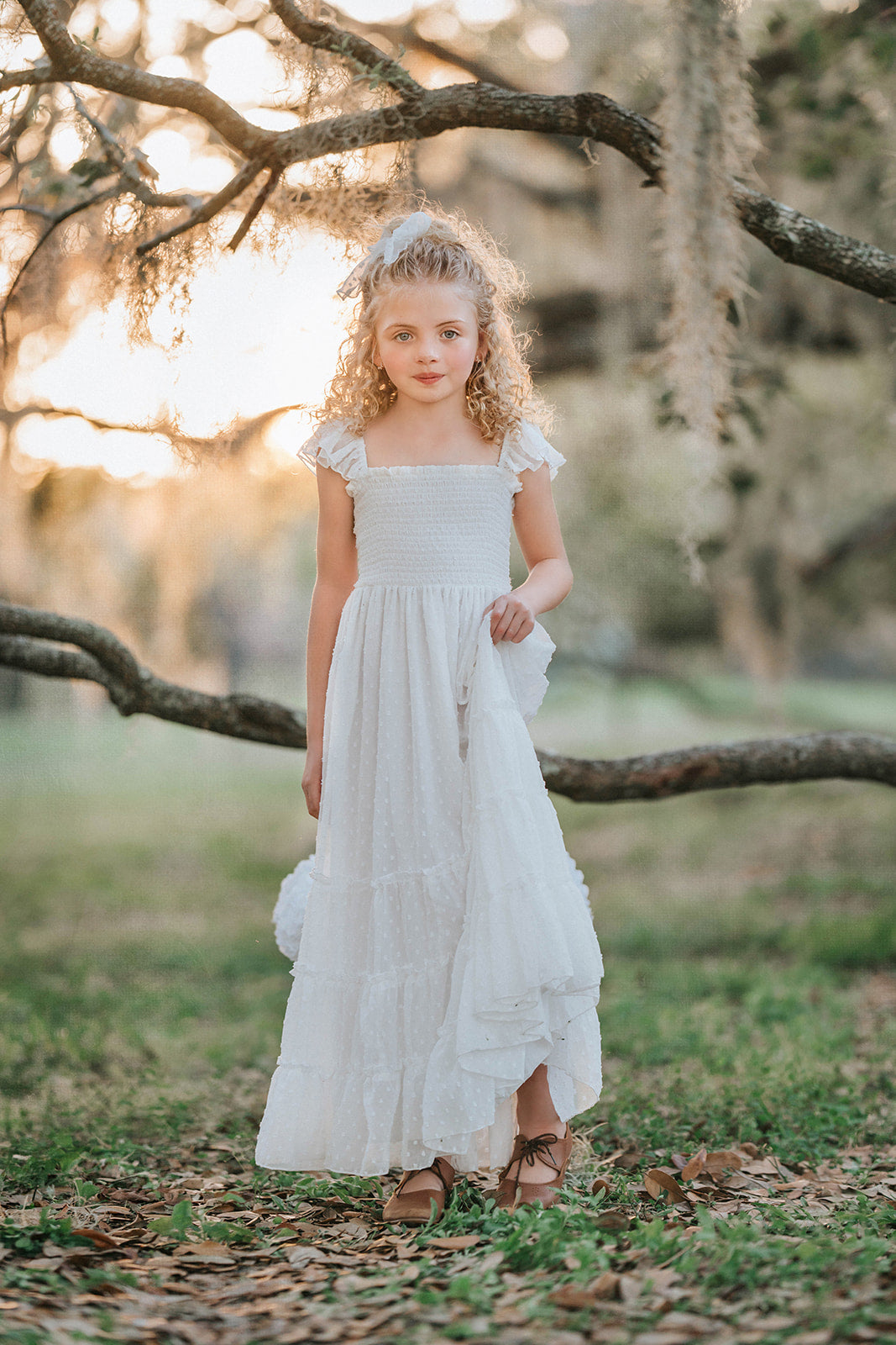 Girl wearing the Orange Blossom Orchard Collection dress in soft white Swiss dot fabric with a smocked bodice, flutter sleeves, and flowing tiered skirt.