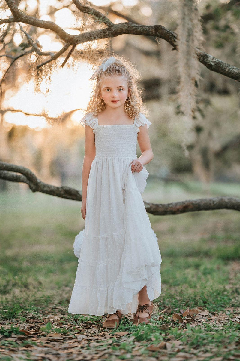 Girl wearing the Orange Blossom Orchard Collection dress in soft white Swiss dot fabric with a smocked bodice, flutter sleeves, and flowing tiered skirt.