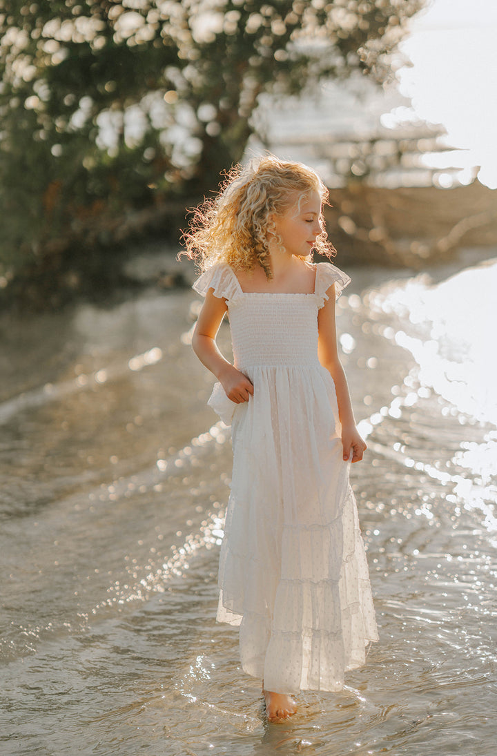 Girl walking barefoot through shallow water wearing the Orange Blossom Orchard Collection dress in soft white Swiss dot fabric.