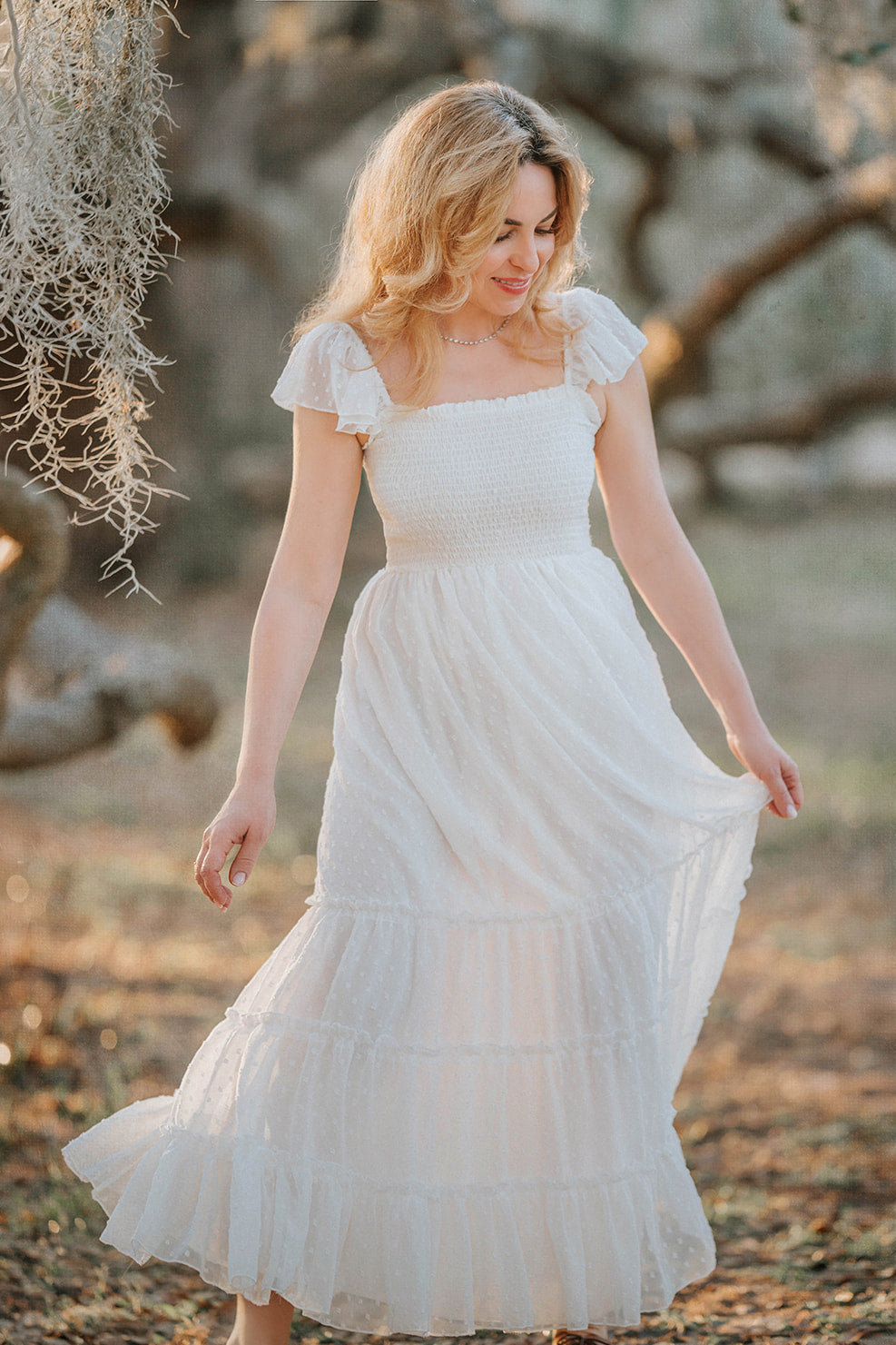 Woman walking through an orchard in the Orange Blossom soft white Swiss dot maxi dress with flutter sleeves.