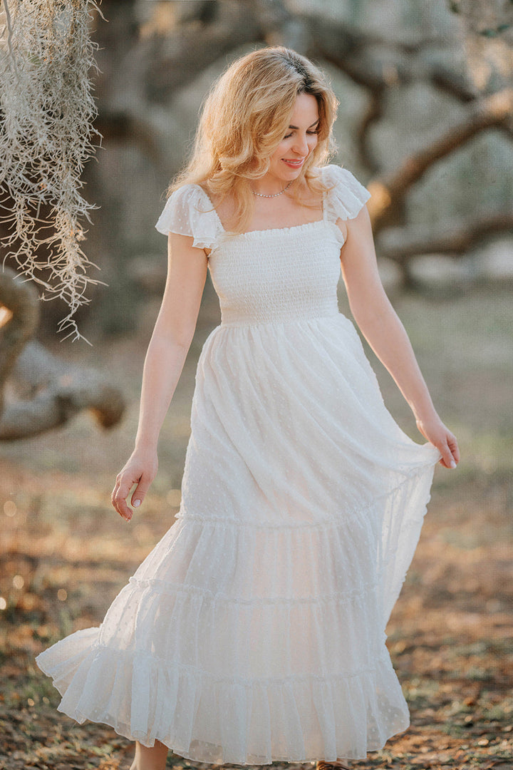 Woman walking through an orchard in the Orange Blossom soft white Swiss dot maxi dress with flutter sleeves.