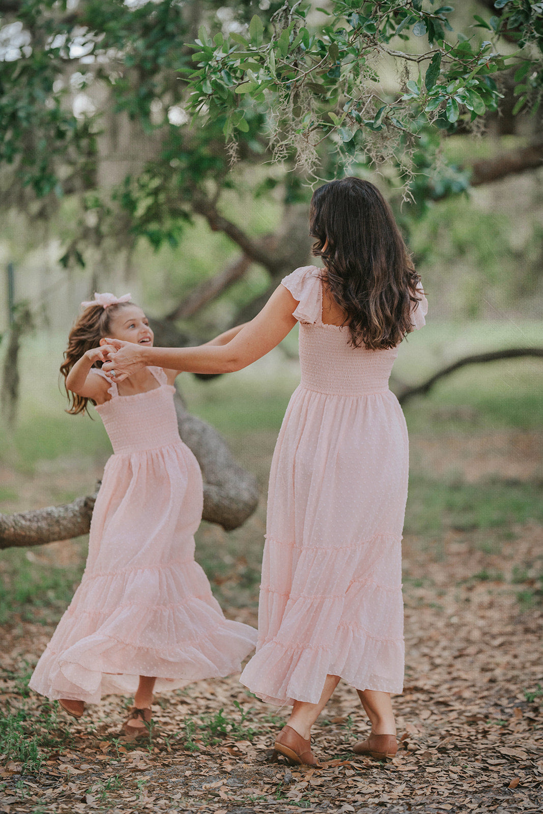 Mother and daughter twirling in matching Wild Strawberry Orchard Collection dresses beneath oak trees.