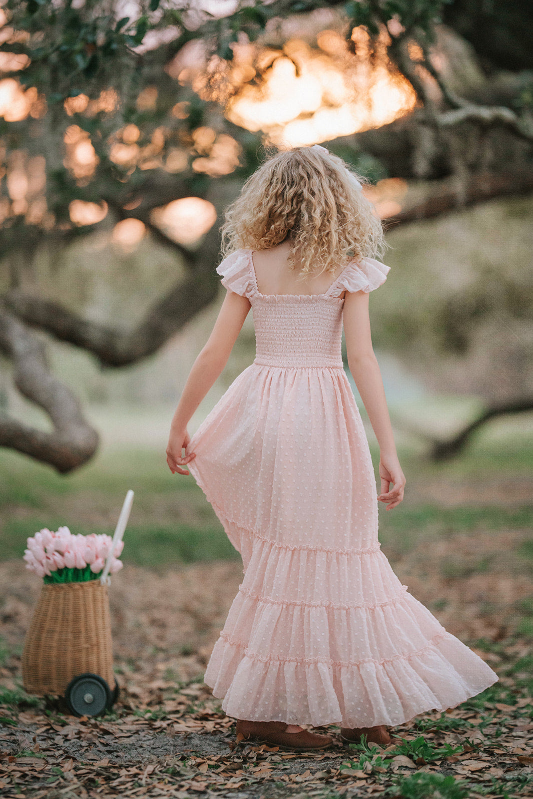 Girl wearing Wild Strawberry Swiss dot dress standing beside flower basket in an orchard